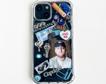 May include: A clear phone case decorated with various charms including a cigarette, a blue gummy bear, a silver chain, a heart-shaped candy with the text HUG ME, a silver key, a portrait of Mac Miller, and the word Capricorn in silver script.
