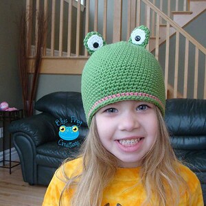 May include: A young girl wearing a green crocheted frog hat with white eyes and pink trim. The hat has large ears and a smiling expression.