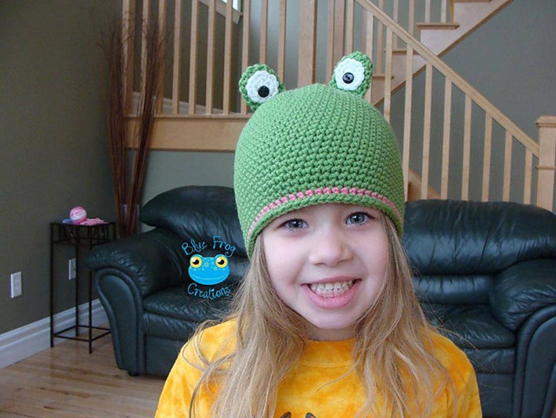 May include: A young girl wearing a green crocheted frog hat with white eyes and pink trim. The hat has large ears and a smiling expression.