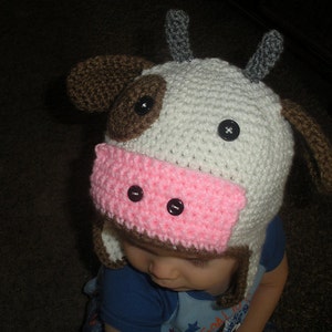 May include: A white and brown crocheted cow hat with pink ears and black button eyes. The hat is worn by a child.