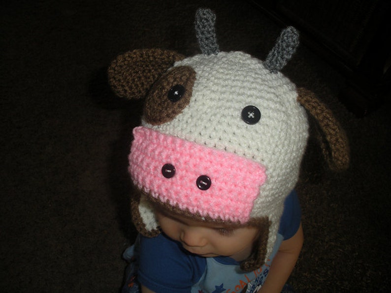 May include: A white and brown crocheted cow hat with pink ears and black button eyes. The hat is worn by a child.