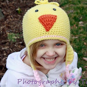 May include: A young girl wearing a yellow crocheted chick hat with a pink and orange beak. The hat has a pink and white trim. The girl is smiling and looking at the camera.