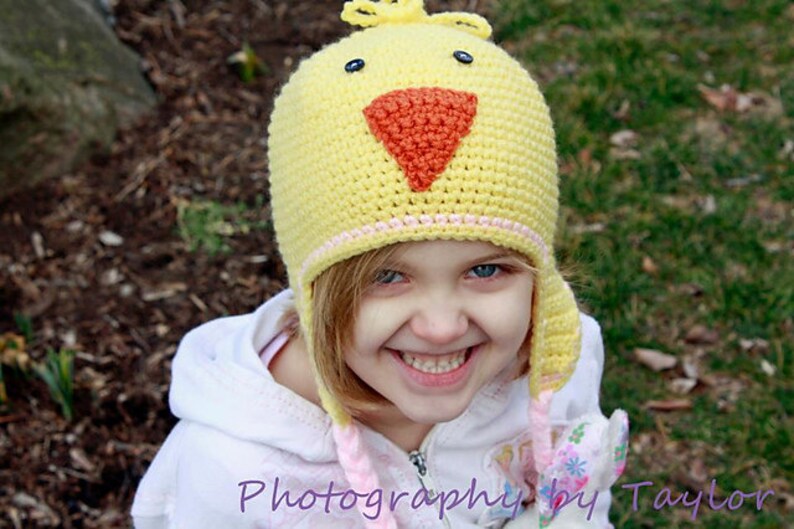 May include: A young girl wearing a yellow crocheted chick hat with a pink and orange beak. The hat has a pink and white trim. The girl is smiling and looking at the camera.