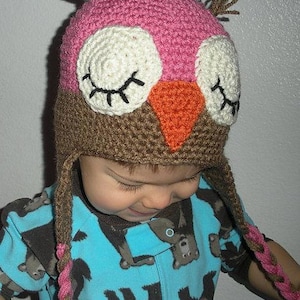 May include: A child wearing a brown and pink crocheted owl hat with long brown and pink braids. The hat has white eyes with black eyelashes and an orange beak.