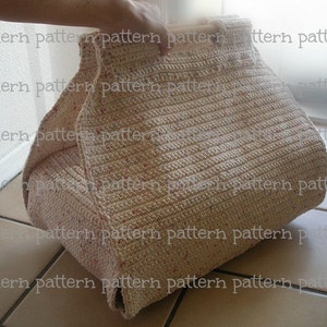 Puede incluir: Una bolsa de mano de crochet con un patrón moteado crema y rosa. La bolsa está doblada y tiene un asa de madera.