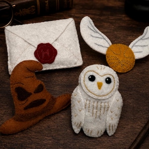 May include: A set of four felt ornaments including a white envelope with a red wax seal, a golden winged ball with stitched details, a brown pointed hat with dark facial features, and a white owl with black eyes and gold embroidered wing details.