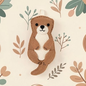 May include: A handmade felt otter ornament in shades of brown and white, with a stitched design. The otter has a white belly, black eyes, and a small, stitched smile. The background features botanical illustrations in muted tones.