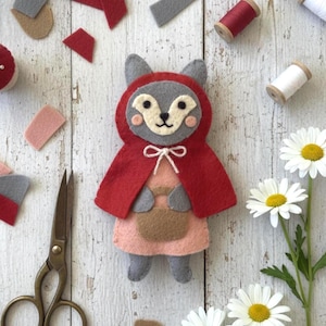 May include: A felt craft project featuring a gray fox character wearing a red hooded cape and a light pink dress. The fox holds a small brown basket and is tied with a white string bow. The figure has stitched facial features and pink cheeks.