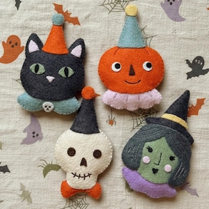 Peut inclure: Quatre décorations en feutrine représentant un chat noir avec un chapeau orange, une citrouille orange avec un chapeau bleu, un crâne blanc avec un chapeau noir et un nœud papillon orange, et une sorcière au visage vert avec un chapeau noir.