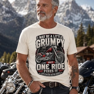 A man with a gray beard and tattoos wears a natural colored t-shirt with a red motorcycle graphic and the text "I MAY BE A LITTLE GRUMPY BUT ONE RIDE FIXES IT LIFE IS BETTER ON TWO WHEELS."