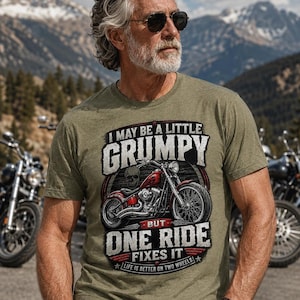 A man with a gray beard, sunglasses, and tattoos wears a heather military green t-shirt with a red motorcycle graphic and the text "I MAY BE A LITTLE GRUMPY BUT ONE RIDE FIXES IT LIFE IS BETTER ON TWO WHEELS."