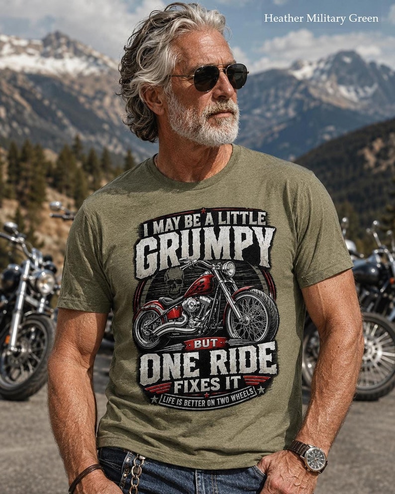 A man with a gray beard, sunglasses, and tattoos wears a heather military green t-shirt with a red motorcycle graphic and the text "I MAY BE A LITTLE GRUMPY BUT ONE RIDE FIXES IT LIFE IS BETTER ON TWO WHEELS."