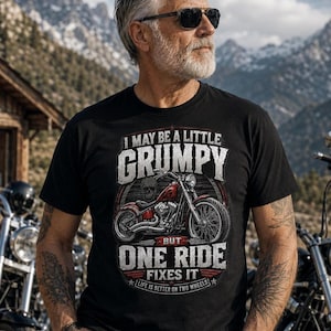 A man with a gray beard, sunglasses, and tattoos wears a black t-shirt with a red motorcycle graphic and the text "I MAY BE A LITTLE GRUMPY BUT ONE RIDE FIXES IT LIFE IS BETTER ON TWO WHEELS."