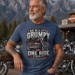A man with a gray beard and tattoos wears an indigo blue t-shirt with a red motorcycle graphic and the text "I MAY BE A LITTLE GRUMPY BUT ONE RIDE FIXES IT LIFE IS BETTER ON TWO WHEELS."