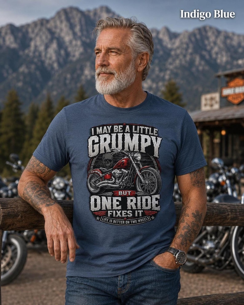 A man with a gray beard and tattoos wears an indigo blue t-shirt with a red motorcycle graphic and the text "I MAY BE A LITTLE GRUMPY BUT ONE RIDE FIXES IT LIFE IS BETTER ON TWO WHEELS."
