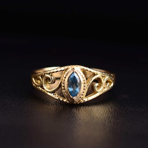 May include: A gold-colored ring with a marquise-cut, light blue gemstone at the center. The ring features an openwork design with intricate scrollwork on the band. The gemstone is set in a detailed bezel.