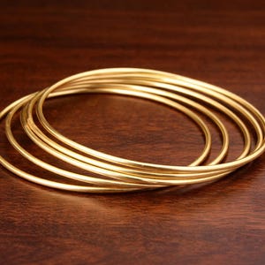 Puede incluir: Un conjunto de seis pulseras doradas, cada una con un diseño simple y redondeado. Las pulseras están apiladas, creando un efecto en capas. Las joyas descansan sobre una superficie de madera marrón oscuro.