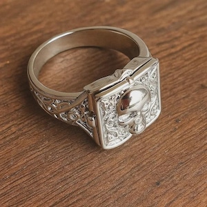 Puede incluir: Un anillo plateado con diseño de calavera. El anillo presenta una cara rectangular adornada con una calavera en relieve y detalles de volutas. La banda tiene un patrón decorativo. El anillo está sobre una superficie de madera.