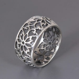 May include: A silver ring with an openwork floral design. The ring features a repeating pattern of stylized flowers and swirling details, creating a delicate and intricate appearance. The ring is shown against a grey background.