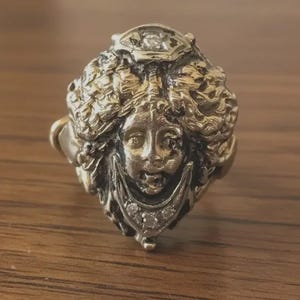 May include: A gold-toned ring featuring a detailed face design with intricate hair and facial features. The ring has a vintage aesthetic, with small, clear stones accenting the face and crown. The ring is set on a gold band.