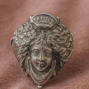 May include: A detailed, silver-toned ring featuring a sculpted face with flowing hair and a crown. The face has delicate features and is set against a textured background. The ring's design is intricate and artistic.