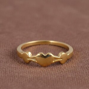 以下が含まれることがあります： 中央にハート型のデザイン、両側に雲のような形が施されたゴールドカラーのリング。茶色の背景に配置され、繊細なディテールが際立っています。