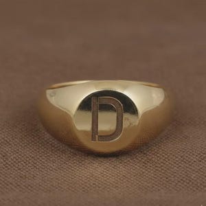 Op de afbeelding: Een goudkleurige zegelring met de letter "D" gegraveerd op een cirkelvormig vlak. De ring heeft een glad, gepolijst oppervlak en een afgeronde vorm. De ring is waarschijnlijk gemaakt van een edelmetaal.