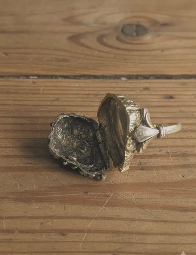 May include: An open, silver-toned locket ring with a detailed, ornate design. The locket is hinged and reveals an interior compartment. The ring features a fleur-de-lis design on the top and a ring band.