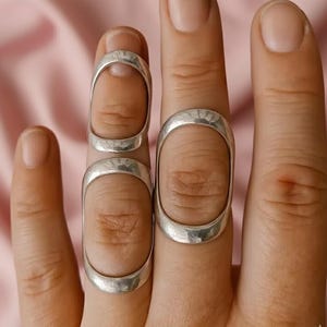 Op de afbeelding: Close-up van drie zilveren vingerringen. De ringen zijn ovaal van vorm en wikkelen zich om de vingers. De ringen hebben een gladde, gepolijste afwerking. De achtergrond is zachtroze.