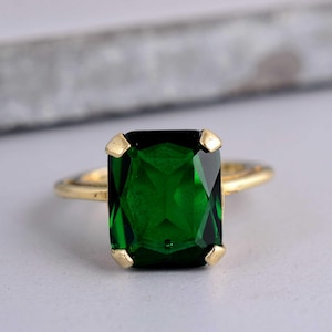 Può includere: Un anello d'oro con una grande pietra verde smeraldo taglio smeraldo. La pietra preziosa è incastonata con quattro griffe d'oro. La fascia dell'anello è in oro sottile e lucido.