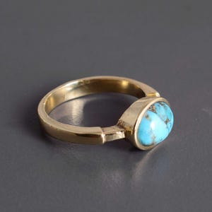 May include: A gold ring with a bright turquoise stone. The ring has a simple band design with a bezel setting that securely holds the oval turquoise gemstone. The ring is displayed against a dark grey background.