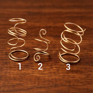 May include: Three gold-colored wire rings in different spiral designs. Each ring is numbered 1, 2, and 3. The rings are displayed on a dark brown wooden surface.
