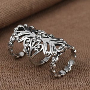 May include: A silver-colored, articulated finger ring with an ornate, floral design. The ring features three bands connected by a decorative, leaf-like centerpiece. The bands have a swirl pattern.
