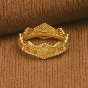 Pode incluir: Um anel dourado com um intrincado design geométrico. O anel apresenta um medalhão central com um emblema detalhado e duas pequenas pedras preciosas transparentes. O anel está sobre um fundo castanho texturizado.