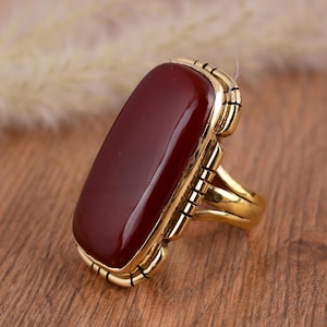 May include: A gold-colored ring featuring a large, rectangular, maroon-colored stone. The ring has a detailed, geometric design around the stone and on the band. The ring is set against a wooden surface.