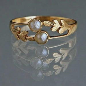 May include: A gold ring with two off-white pearls. The ring has a leaf design on the sides and around the pearls. The ring is reflected on a shiny surface. The ring's design includes leaf shapes.