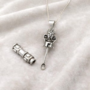 May include: A silver pendant necklace with an intricate design. The pendant has a stylized figure with a long, spoon-like element. A separate, cylindrical component is also visible, possibly an accessory. The necklace is displayed on a soft, white surface, likely fabric.