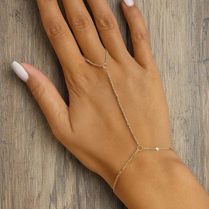 Puede incluir: Una delicada pulsera de cadena de mano dorada. La joya se conecta al dedo anular y a la muñeca con una fina cadena. La pulsera presenta pequeños detalles circulares y en forma de corazón. La mano tiene esmalte de uñas blanco.