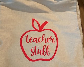 Può includere: Una borsa tote in tela beige con manici lunghi. Una grafica di mela rossa con le parole "Teacher Stuff" è stampata sul davanti. La borsa è progettata per trasportare libri, forniture o altri oggetti.