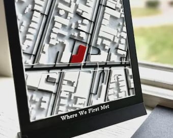 May include: A square black frame containing a 3D white street map with a single red building highlighted. The frame features the text Where We First Met. Beside it lies a black square plaque with the text Joyful Little Prints.