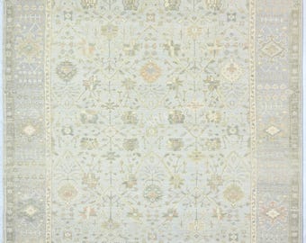 May include: A rectangular area rug featuring a light gray and cream background with a repeating floral and geometric pattern in muted tones of sage green, soft peach, and beige. The rug has a wide border with intricate linear motifs and fringe.