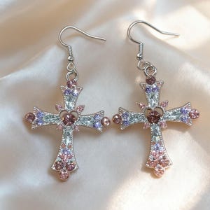 May include: A pair of silver-toned cross earrings. Each cross is embellished with pink, purple, and clear crystals. The earrings have a hook closure.