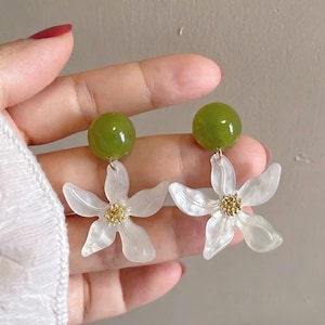 May include: A pair of floral earrings. Each earring features a translucent white flower with a gold-colored center and a green, spherical top. The earrings are designed to dangle and are shown against a neutral background.