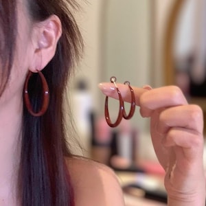 Może przedstawiać: Ciemnoczerwone kolczyki koła. Jeden jest noszony na uchu, drugi trzymany przez palce. Kolczyki mają gładkie, błyszczące wykończenie i kształt łzy.