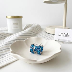 Puede incluir: Pendientes de aro plateados con gemas azules cuadradas y facetadas. Se exhiben en un plato blanco con forma de pétalo. Una tarjeta indica "THE BLUE COLLECTION".