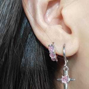 Puede incluir: Primer plano de una oreja adornada con dos pendientes de plata. Uno es un aro pequeño con tres gemas rosas, y el otro es una cruz colgante con una gema rosa en el centro. Los pendientes se colocan contra el pelo oscuro.