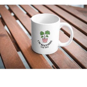 Może przedstawiać: Biały ceramiczny kubek z ilustracją zielonej rośliny w różowej doniczce. Kubek ma napis "I'm rooting for you" w zakrzywionej czcionce. Kubek stoi na brązowej drewnianej powierzchni.
