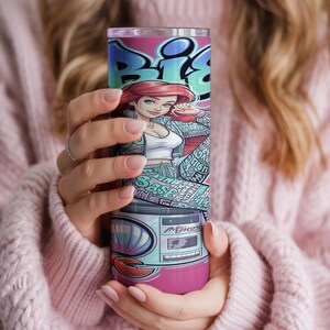 Pu&ograve; includere: Un bicchiere cilindrico in acciaio inossidabile con un vivace design in stile graffiti. L'illustrazione mostra un personaggio dei cartoni animati con capelli rossi, un top bianco e una giacca fantasia, sopra una grafica di un boombox retr&ograve; con conchiglia.