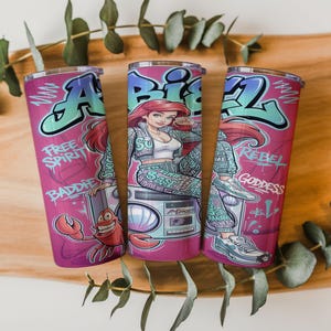 Pu&ograve; includere: Tre bicchieri cilindrici con sfondo rosa raffiguranti un personaggio dei cartoni animati con capelli rossi, un granchio e un boombox. Il design include scritte in stile graffiti Ariel, Free Spirit, Baddie, Rebel e Goddess in blu e bianco.