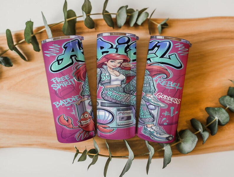 Pu&ograve; includere: Tre bicchieri cilindrici con sfondo rosa raffiguranti un personaggio dei cartoni animati con capelli rossi, un granchio e un boombox. Il design include scritte in stile graffiti Ariel, Free Spirit, Baddie, Rebel e Goddess in blu e bianco.